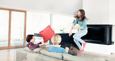 Eltern auf Couch, Tochter springt, rotes Polster fliegt in der Luft